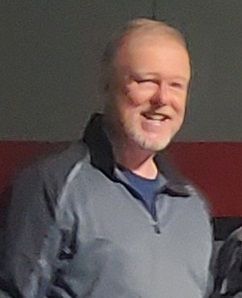 A man with a beard is smiling and wearing a gray jacket.
