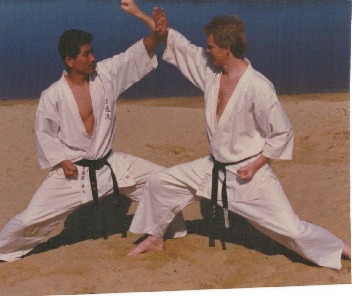 Two men in white karate uniforms are kneeling on the beach