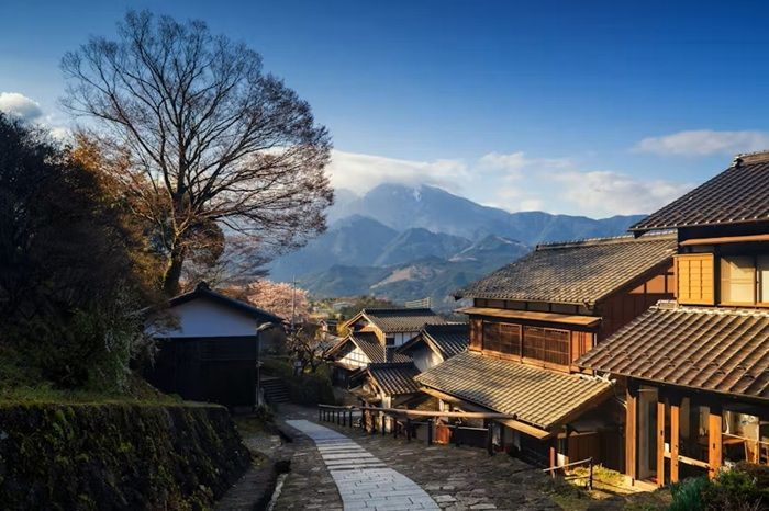 Stone path in Japanese village with traditional buildings, mountains, and blue sky.
