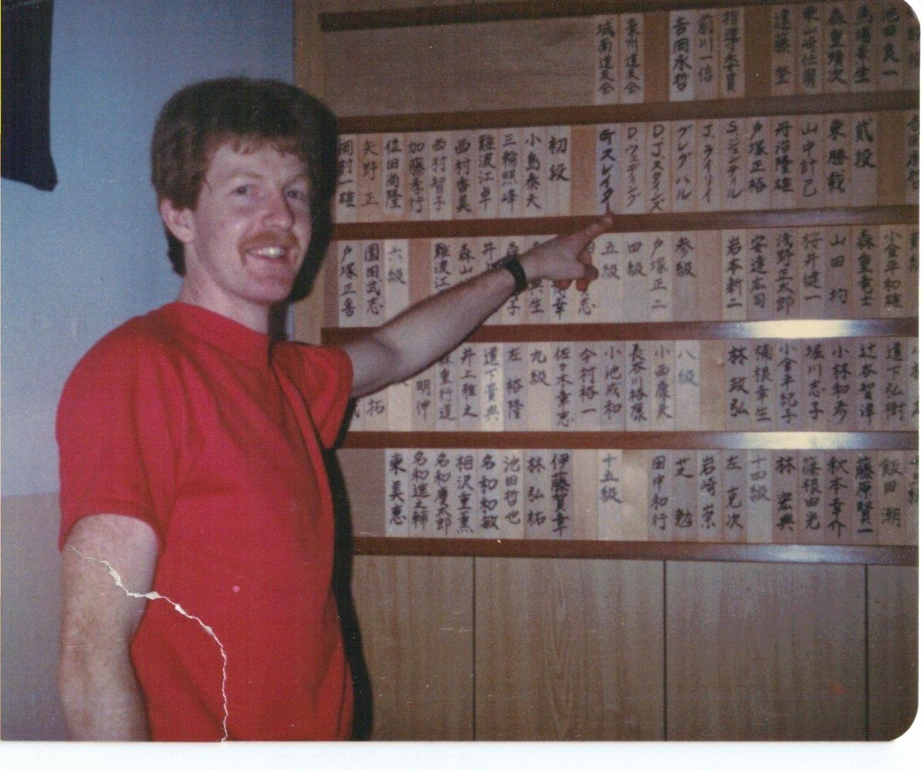 A man in a red shirt is pointing at a wall with chinese writing on it