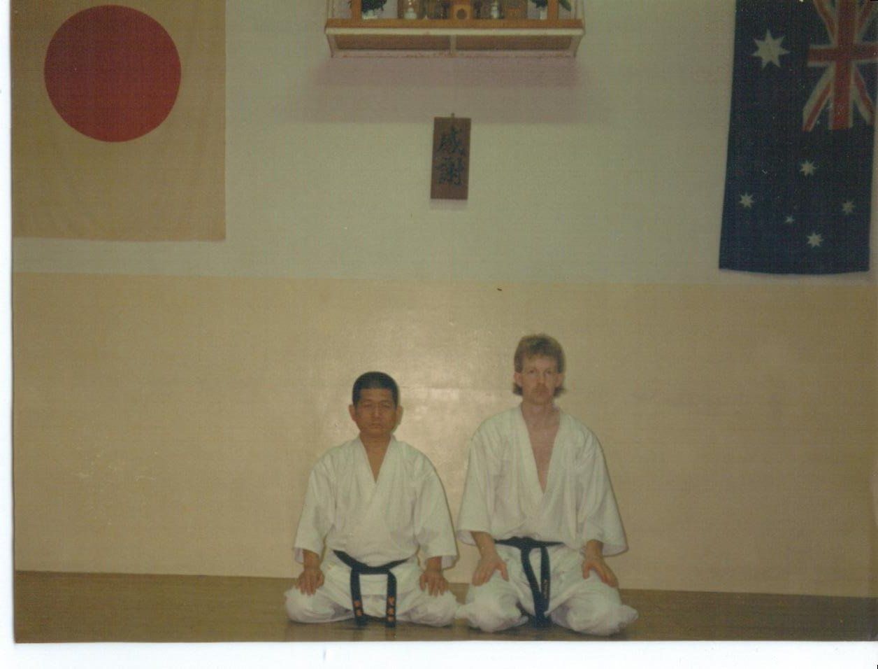 Two men are sitting on the floor in front of a wall with flags on it.