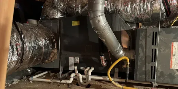 A furnace with ductwork in an enclosed space. The unit has metal vents and pipes. Yellow gas line attached.