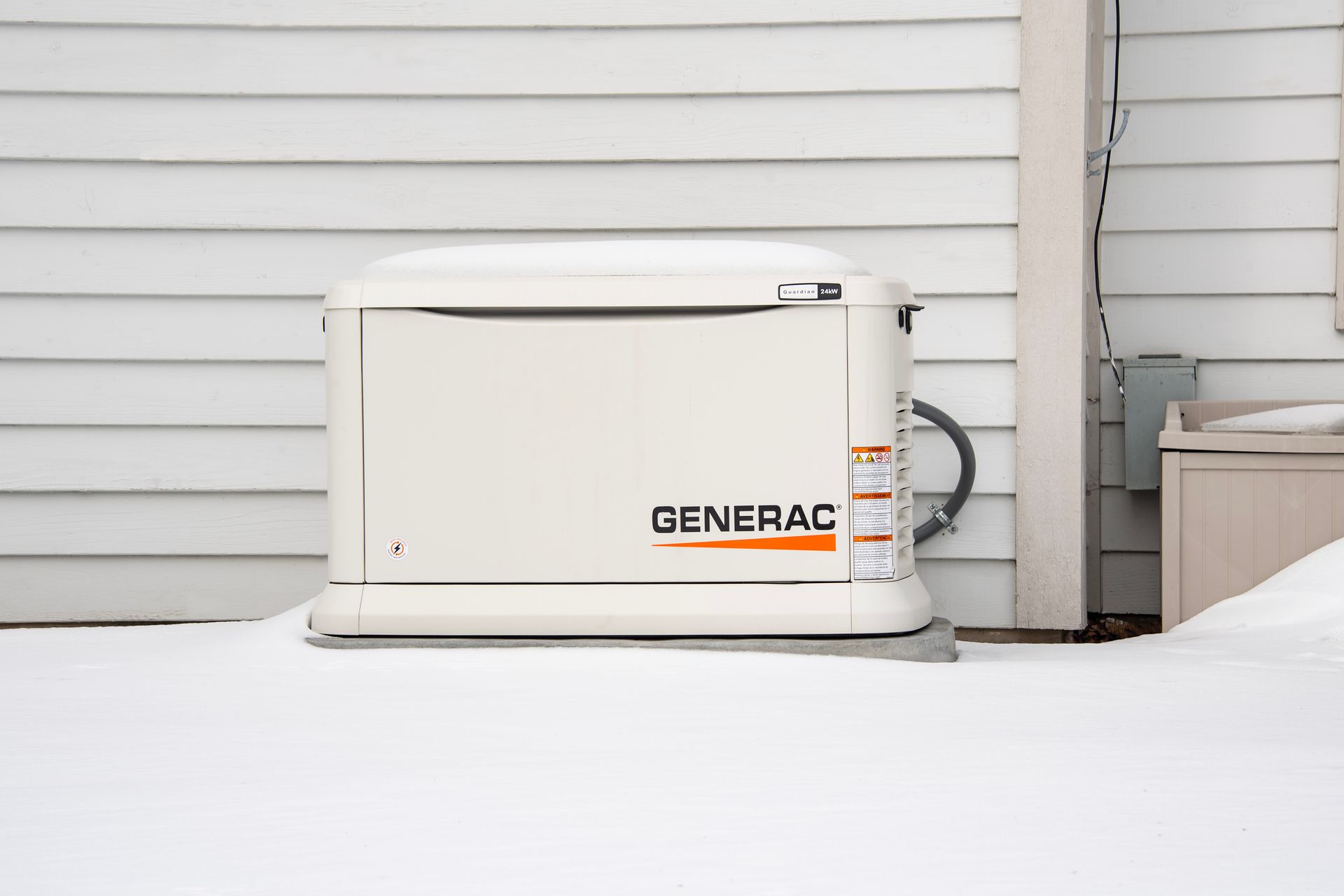 A Generac generator, off-white, sits in snow against a white-sided house, in an outdoor setting.