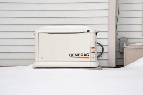 A Generac generator, off-white, sits in snow against a white-sided house, in an outdoor setting.