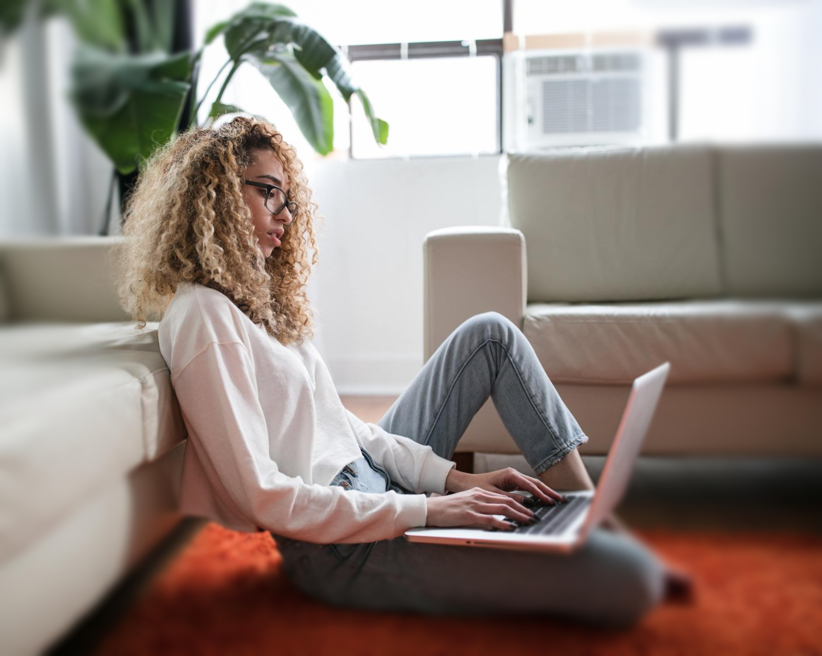 A young woman sitting on an orange rug looking at her laptop