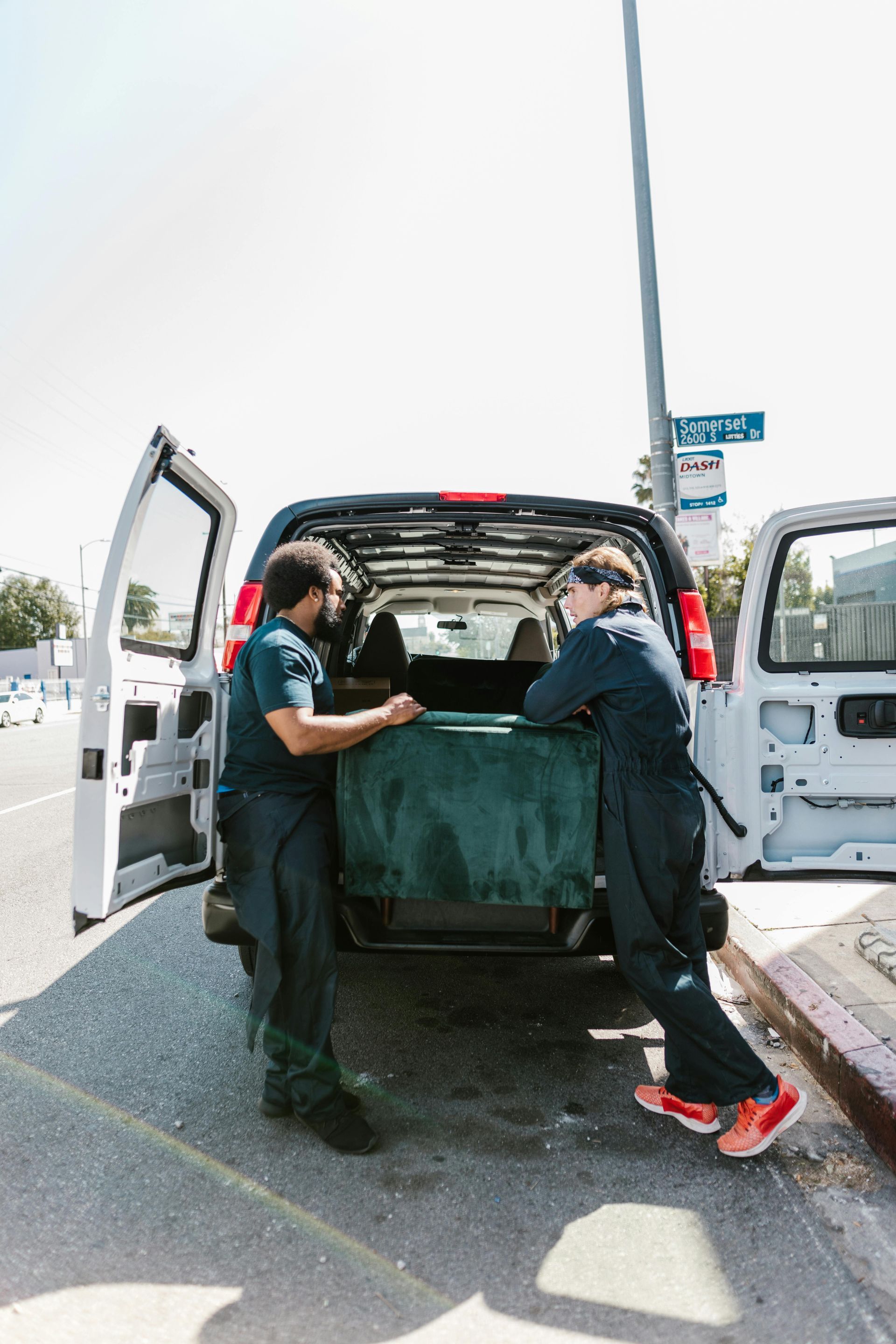 Two workers leaning on a van open in a sunny urban setting. One looks at a teal upholstered object.
