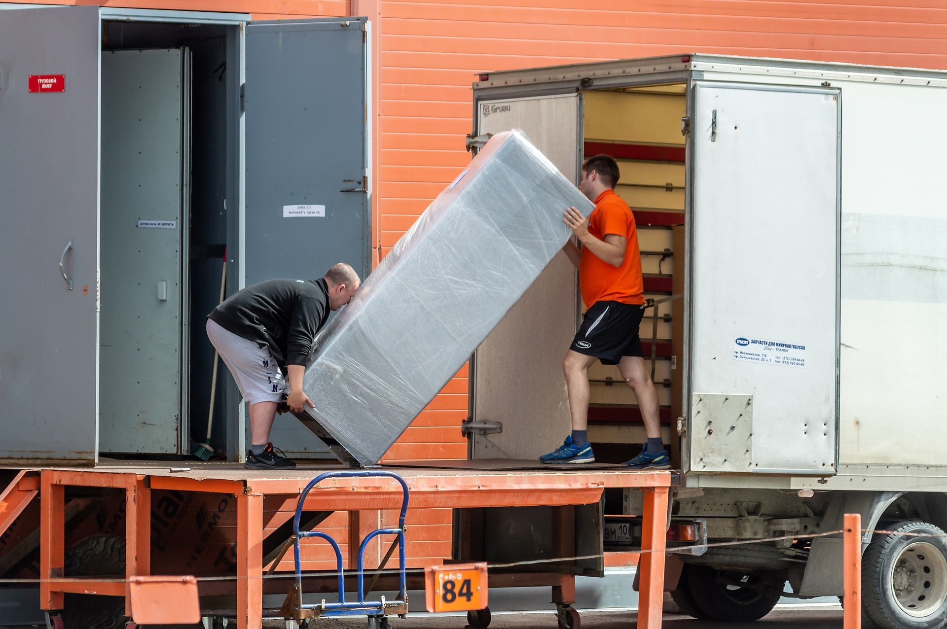 Two people loading a wrapped mattress into a truck from a loading dock.