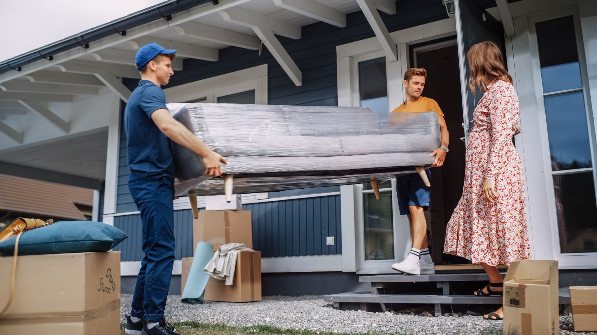 Movers carrying a couch wrapped in plastic into a house; a woman watches.