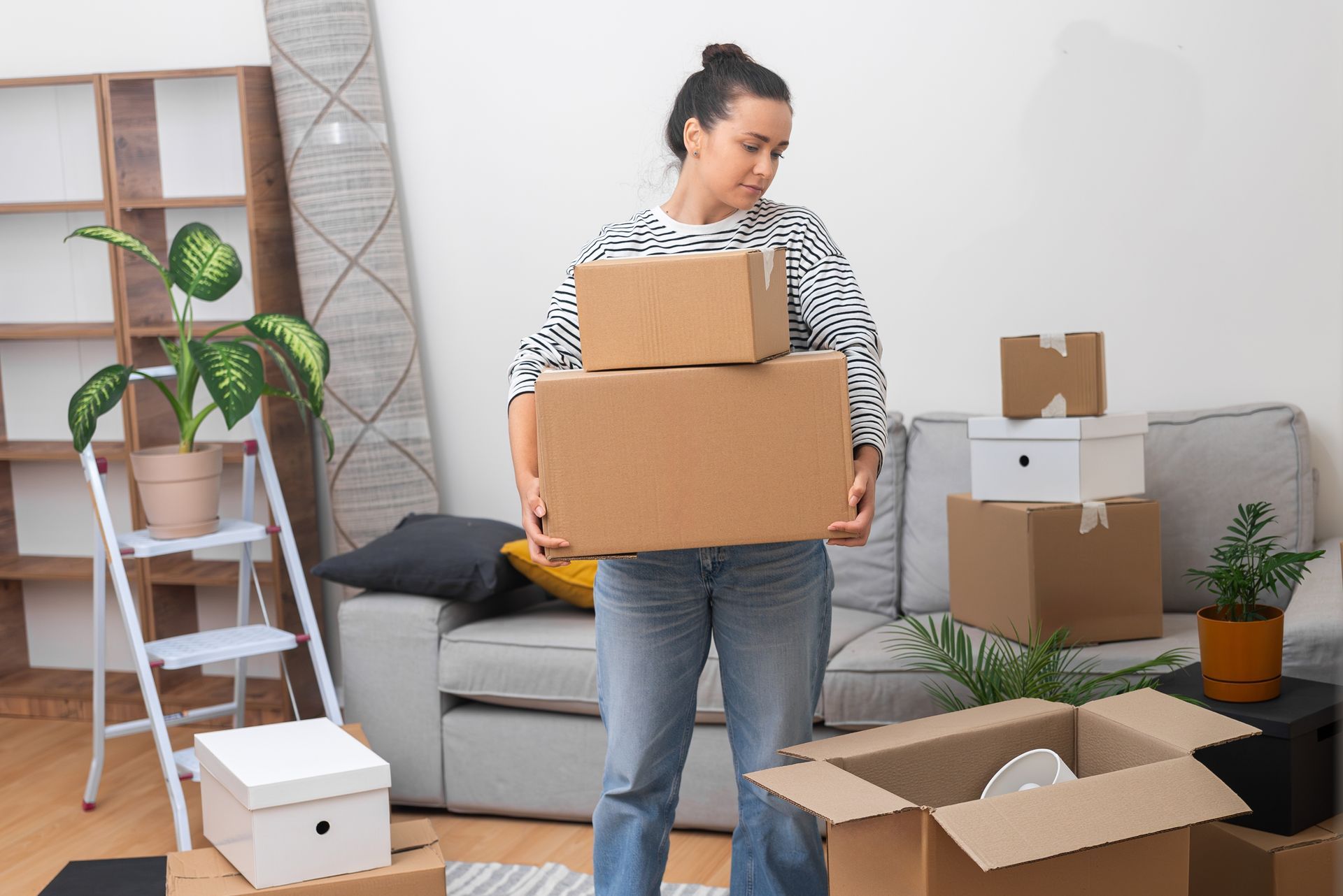 Woman holding boxes indoors, looks down. Boxes, couch, plant, and other packed boxes are visible.