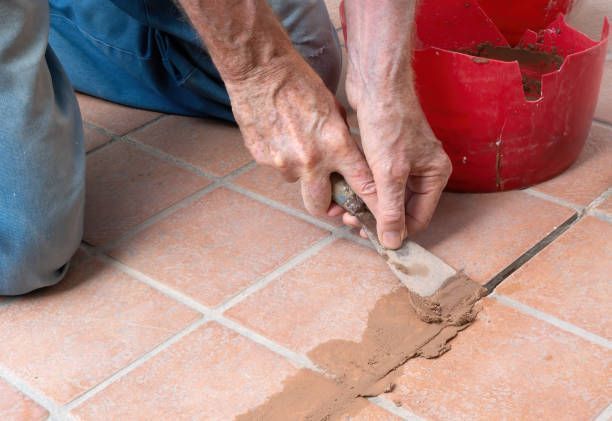 A person is kneeling on the floor using a spatula to repair a tile floor.