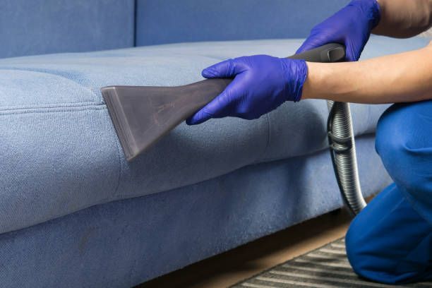 A person is cleaning a blue couch with a vacuum cleaner.
