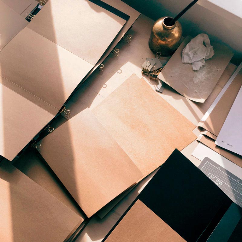 piles of kraft brown paper sit on a desk, in various stages of being constructed into notebooks