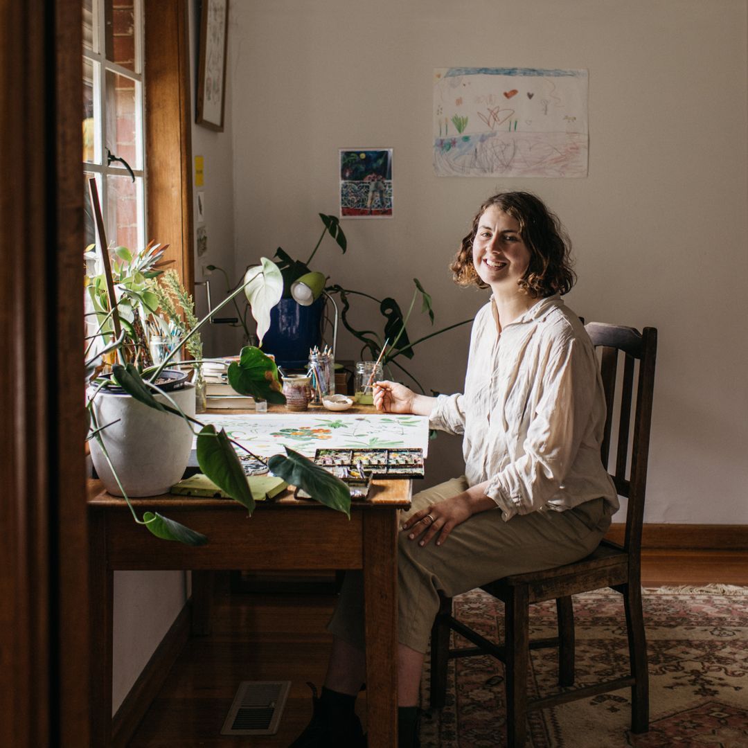 Claire Mosley sits at a wooden desk, working on a watercolour painting of plants
