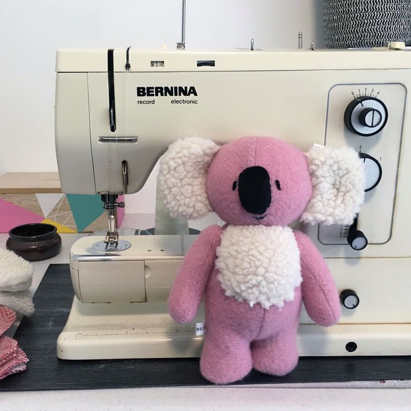 a soft toy koala made from pink fabric with fluffy white ears, propped against a sewing machine