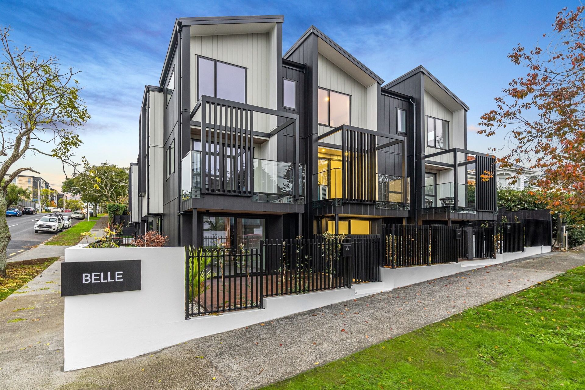 Modern townhouses with dark siding, black railings, and glass balconies on a street with a sidewalk and green lawn.