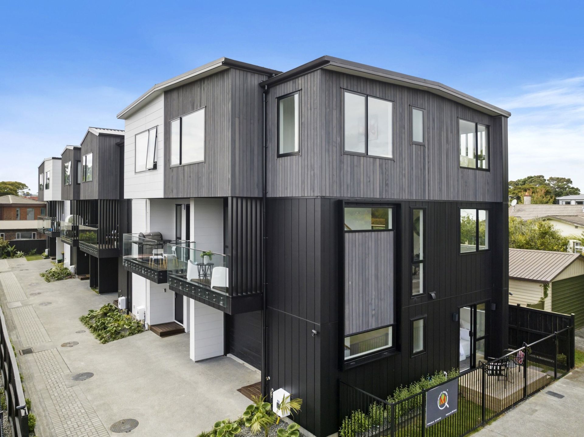 Modern three-story townhouses with dark and light gray siding, glass balconies, and a brick driveway under a blue sky.