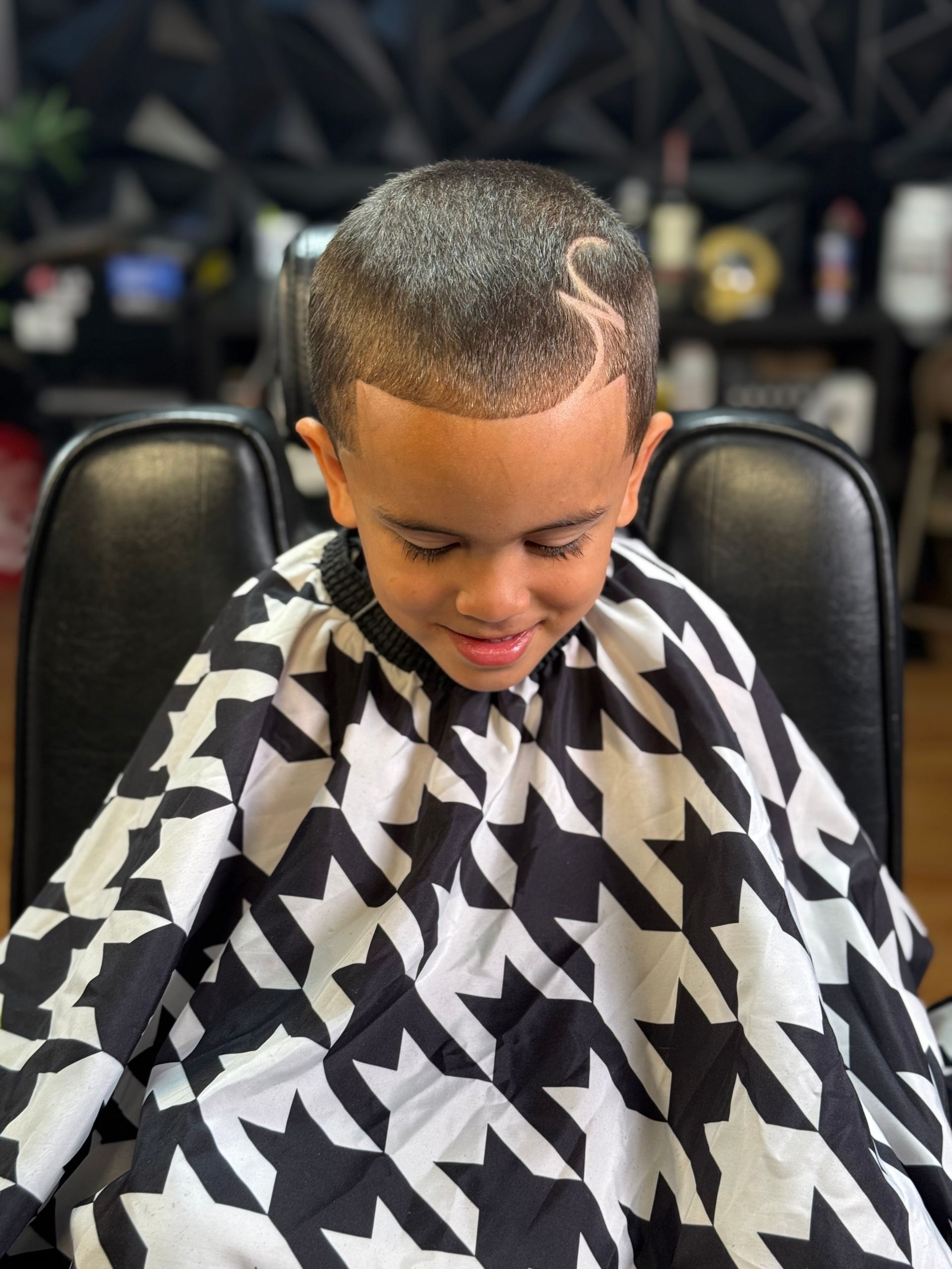 Boy in barber chair with fresh haircut, wearing black and white patterned cape.