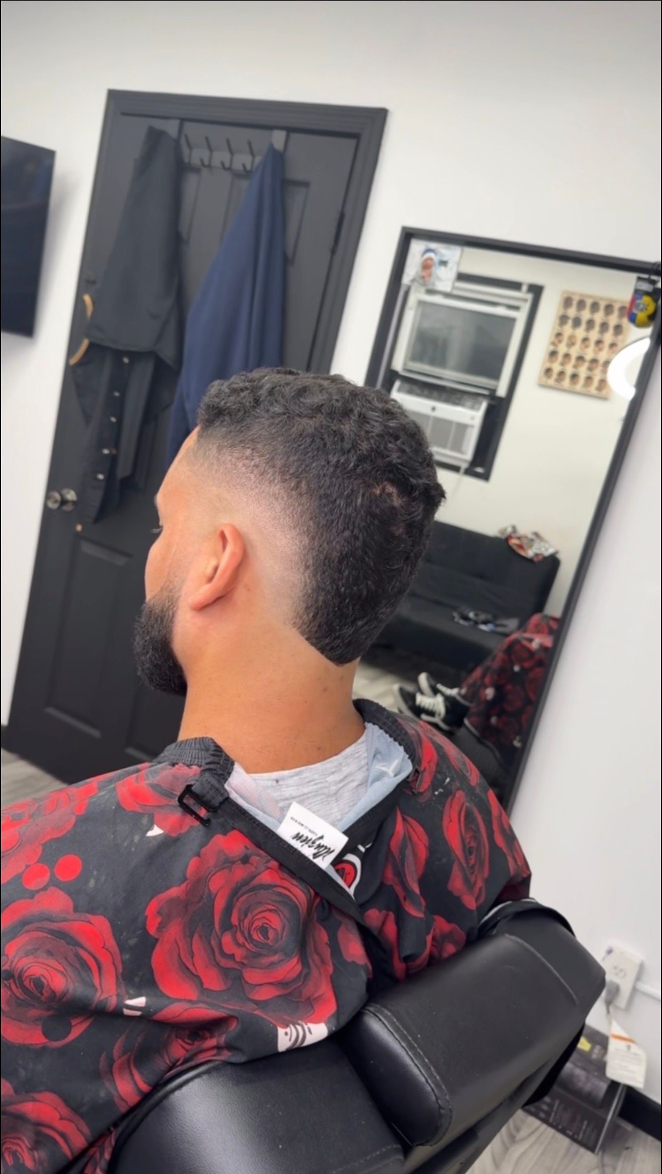 Man getting a haircut at a barbershop. He has a fade with textured top. Black barber chair, mirror.