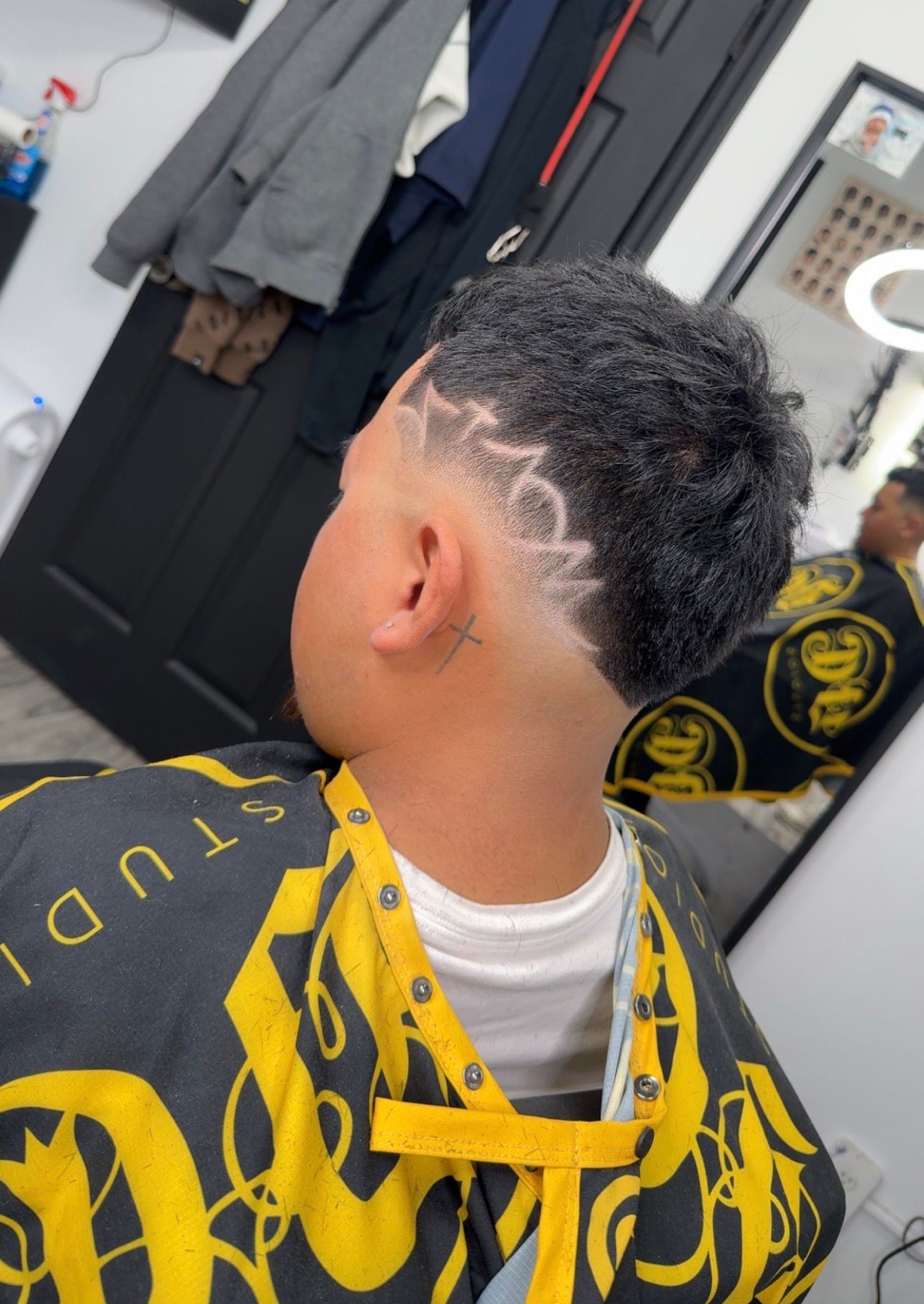 Person with a fade haircut and design on the side of their head; in a barber shop wearing a yellow and black cape.