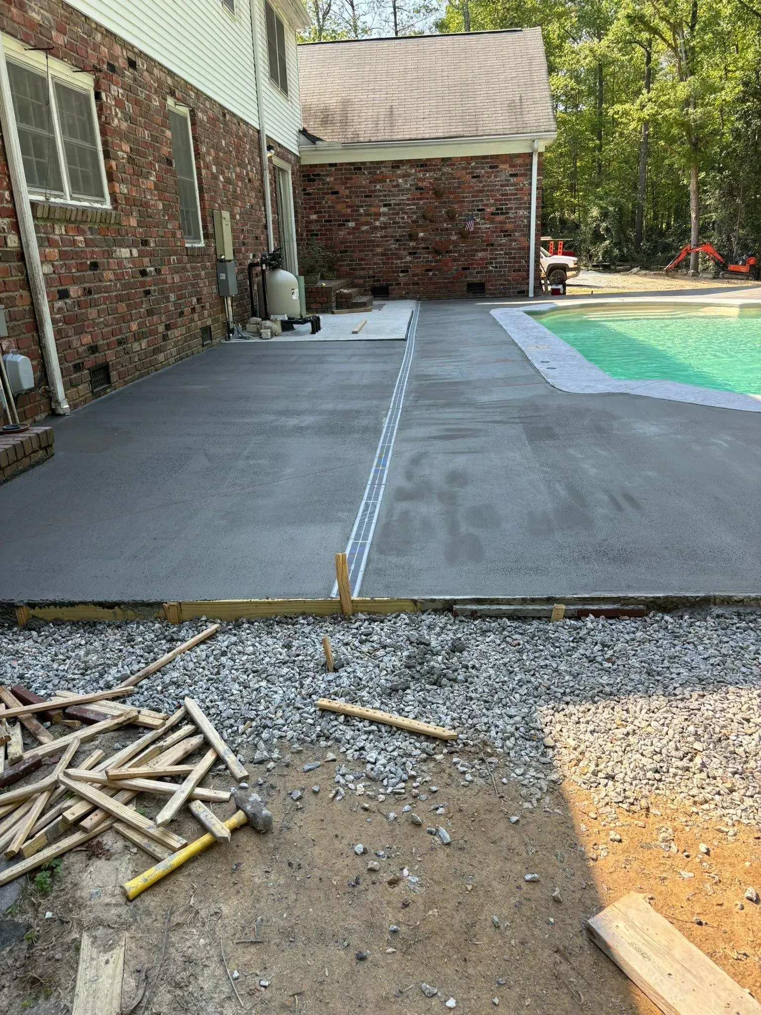 Concrete patio under construction next to a brick house and pool. Gravel, tools visible.