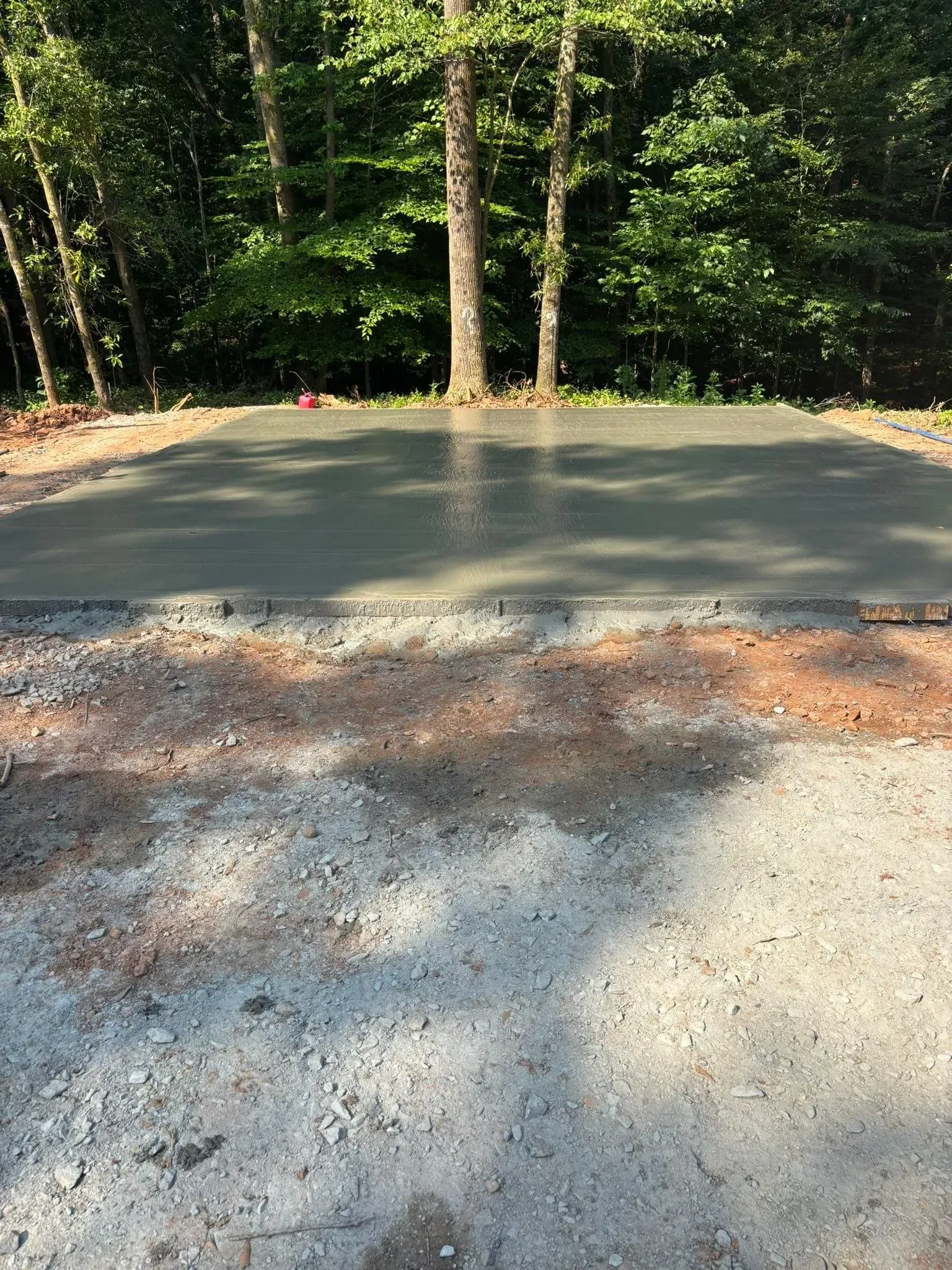 A newly poured concrete pad with gravel and trees in the background.