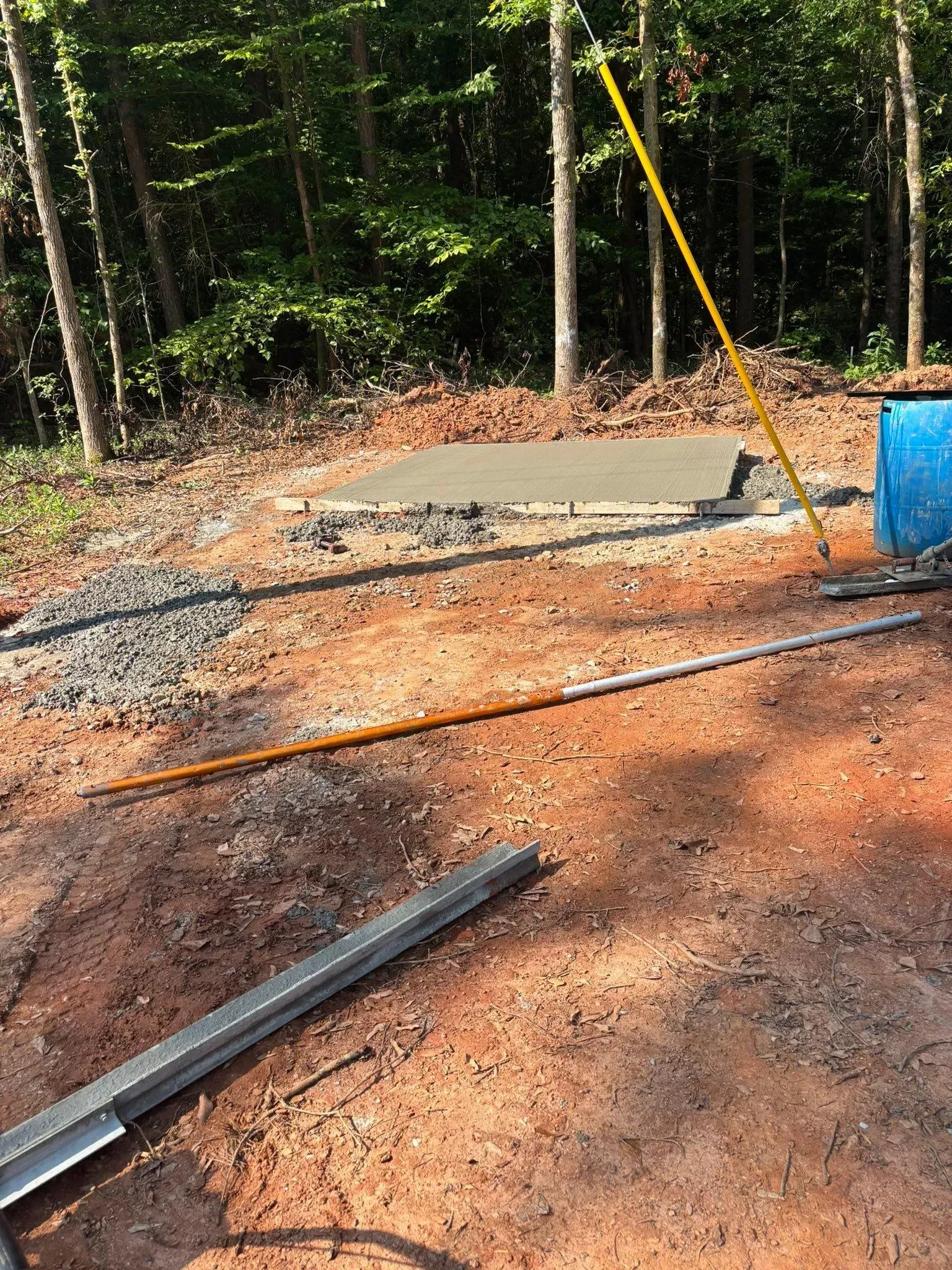 Construction site with concrete slab, tools, and a blue barrel in a wooded area.