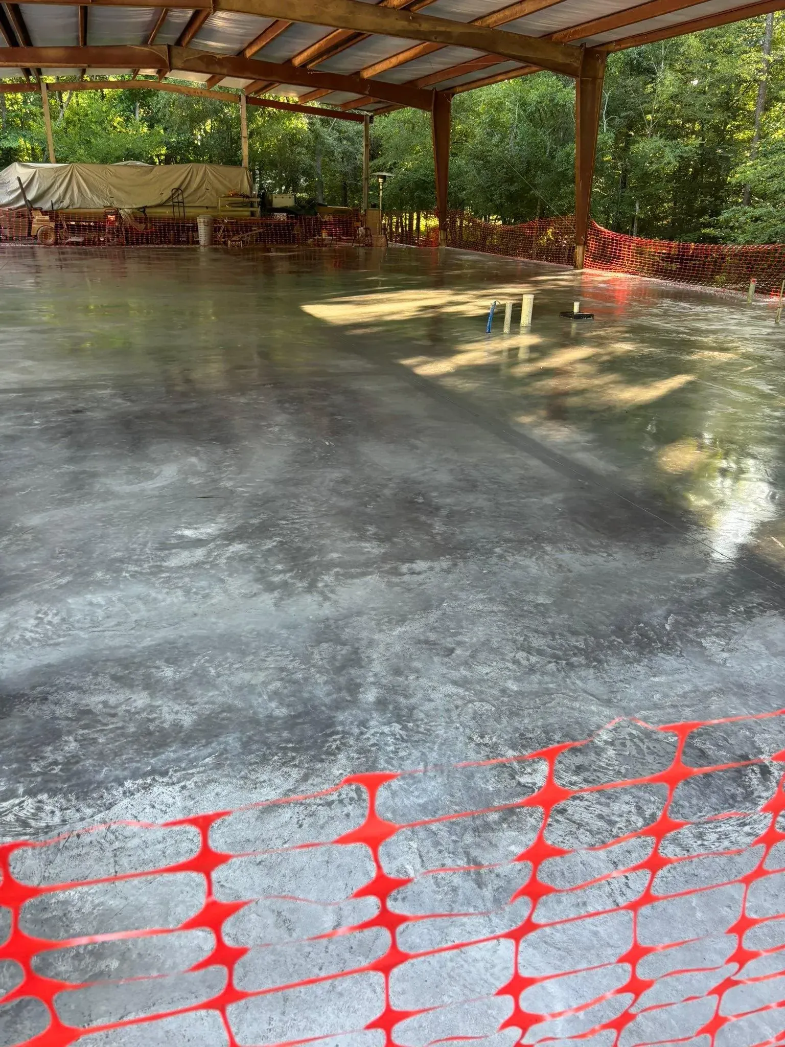 A concrete patio area with a red construction fence, under a wooden structure with trees in the background.