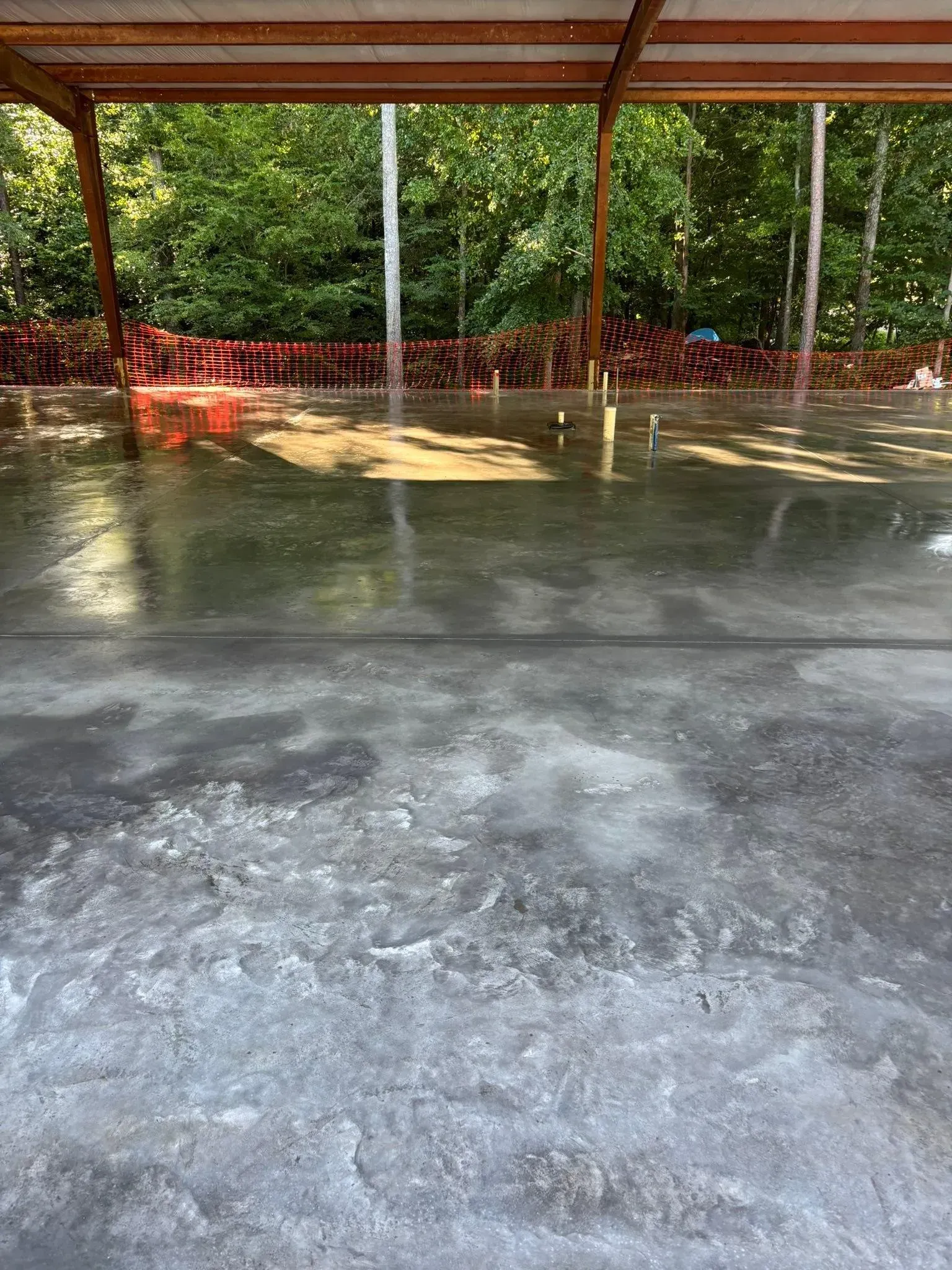 Wet concrete floor under a metal-roofed structure, with trees and stacks of red items in the background.