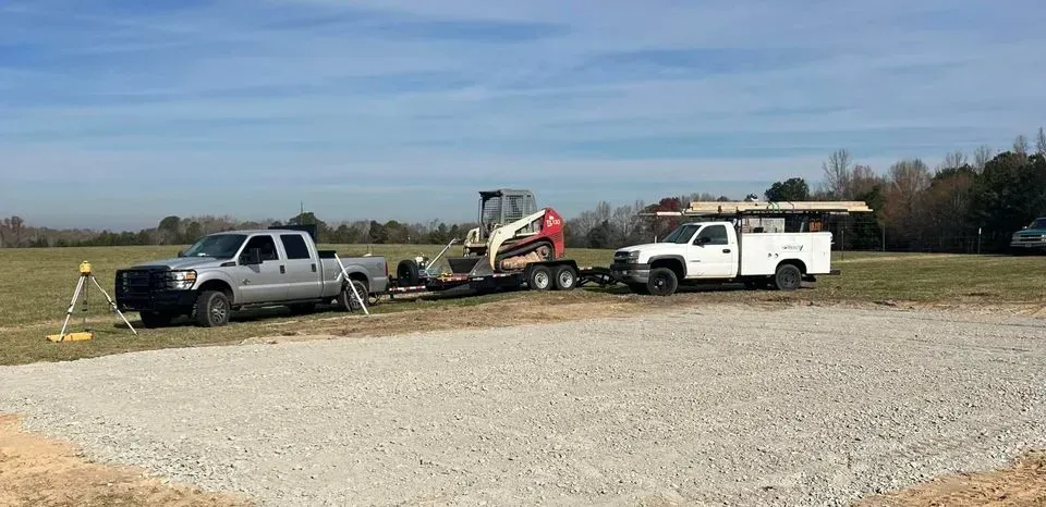 Two trucks and a Bobcat on a trailer parked on gravel in a field on a clear day.