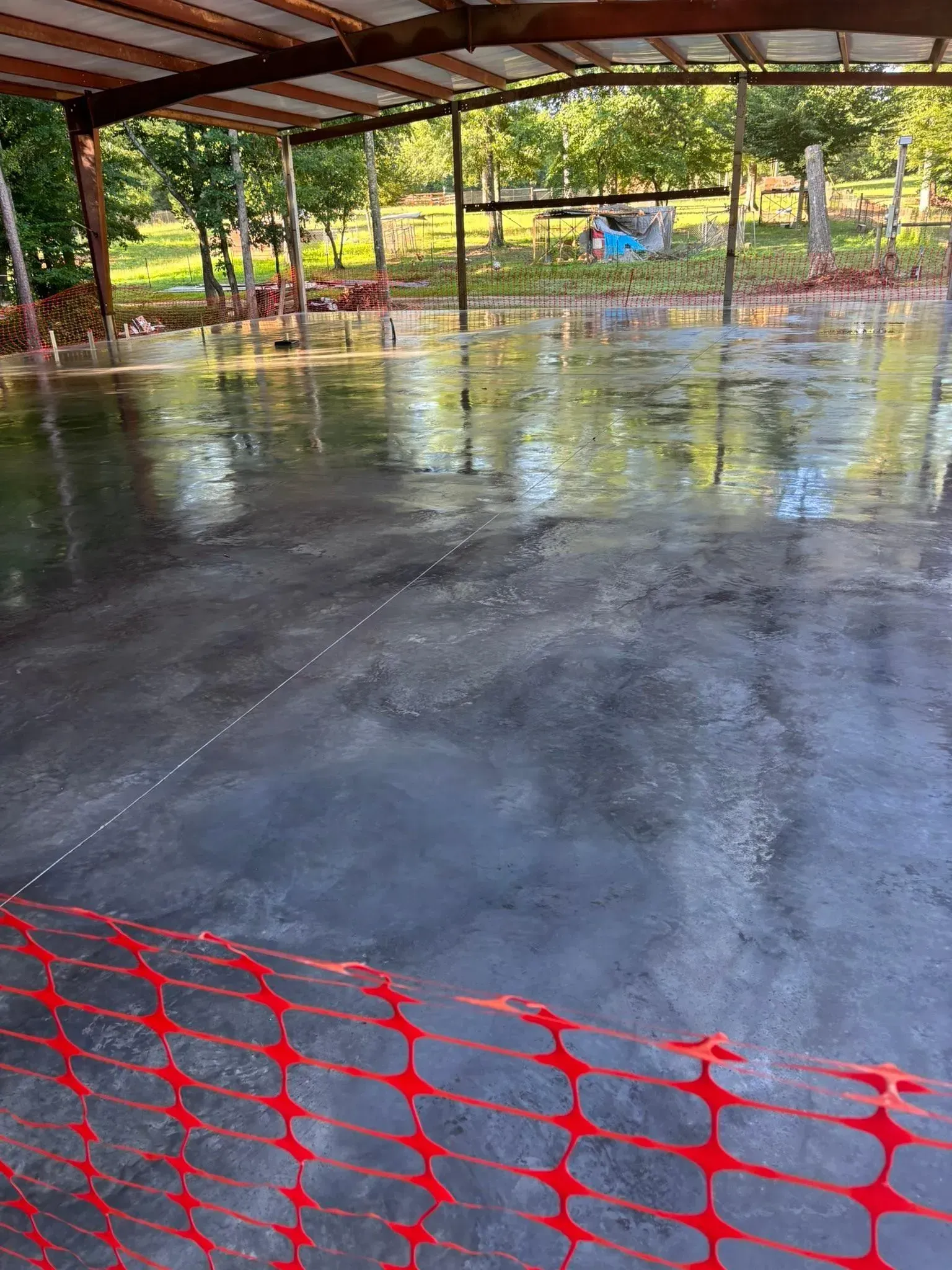 Newly poured wet concrete floor under a metal roof, with orange safety fencing and trees in the background.