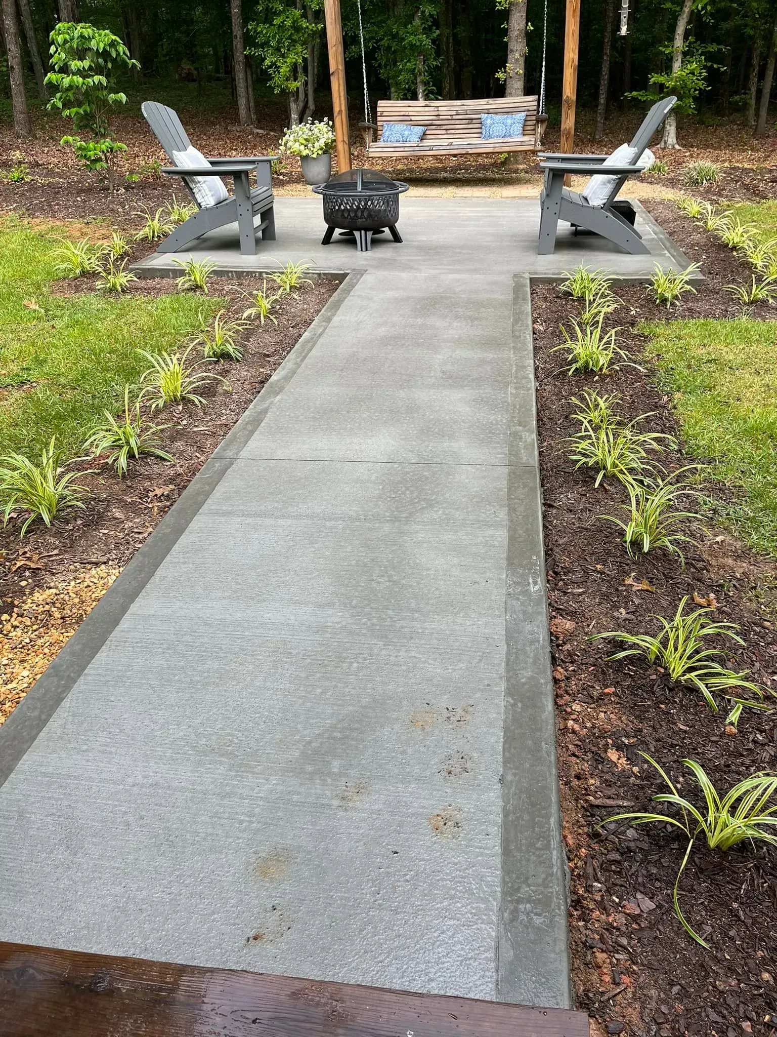 Concrete pathway leads to a fire pit with two chairs, a wooden structure, and garden beds in a backyard.