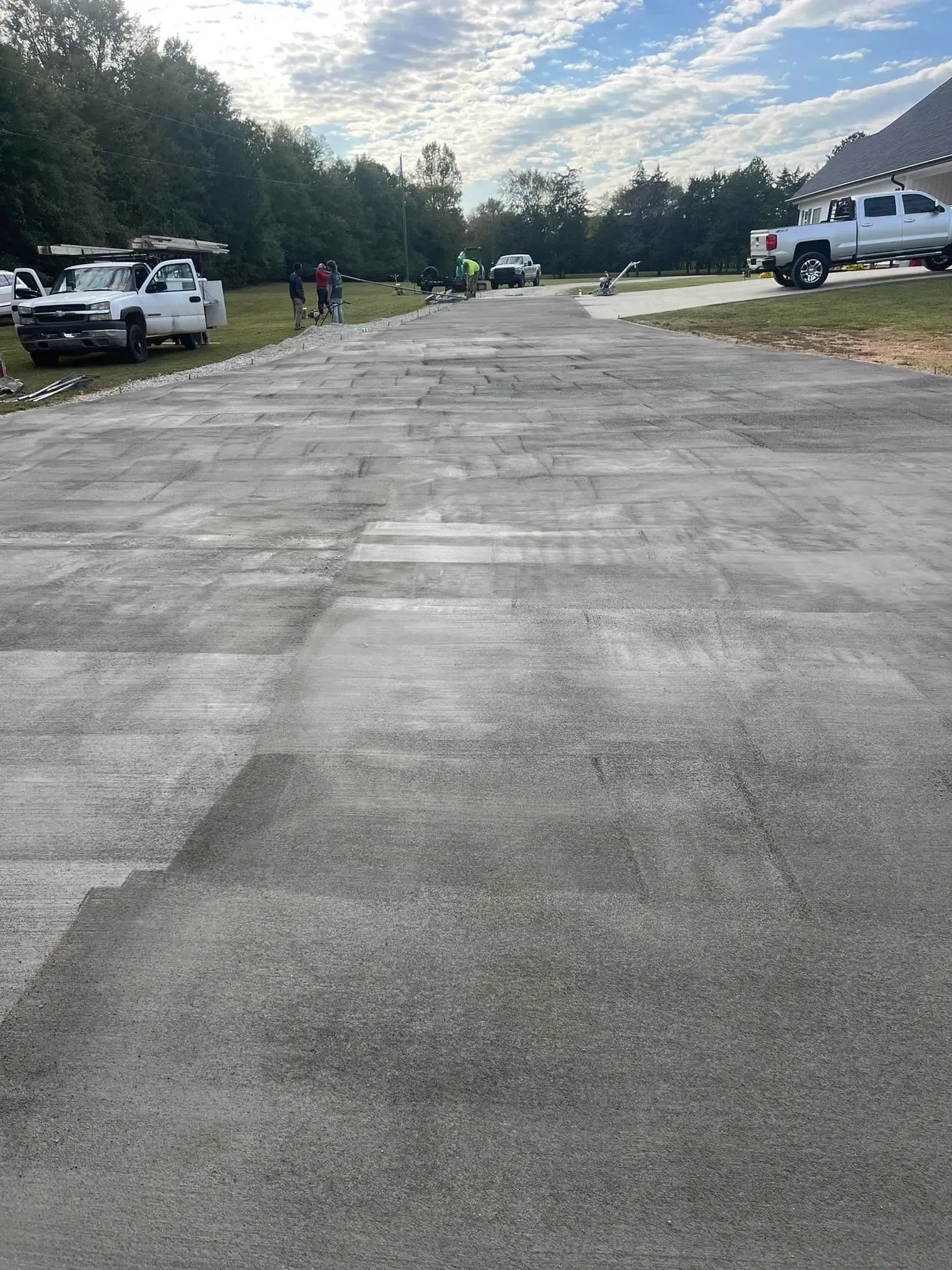 Workers on a newly paved concrete surface, with parked trucks and trees under a cloudy sky.