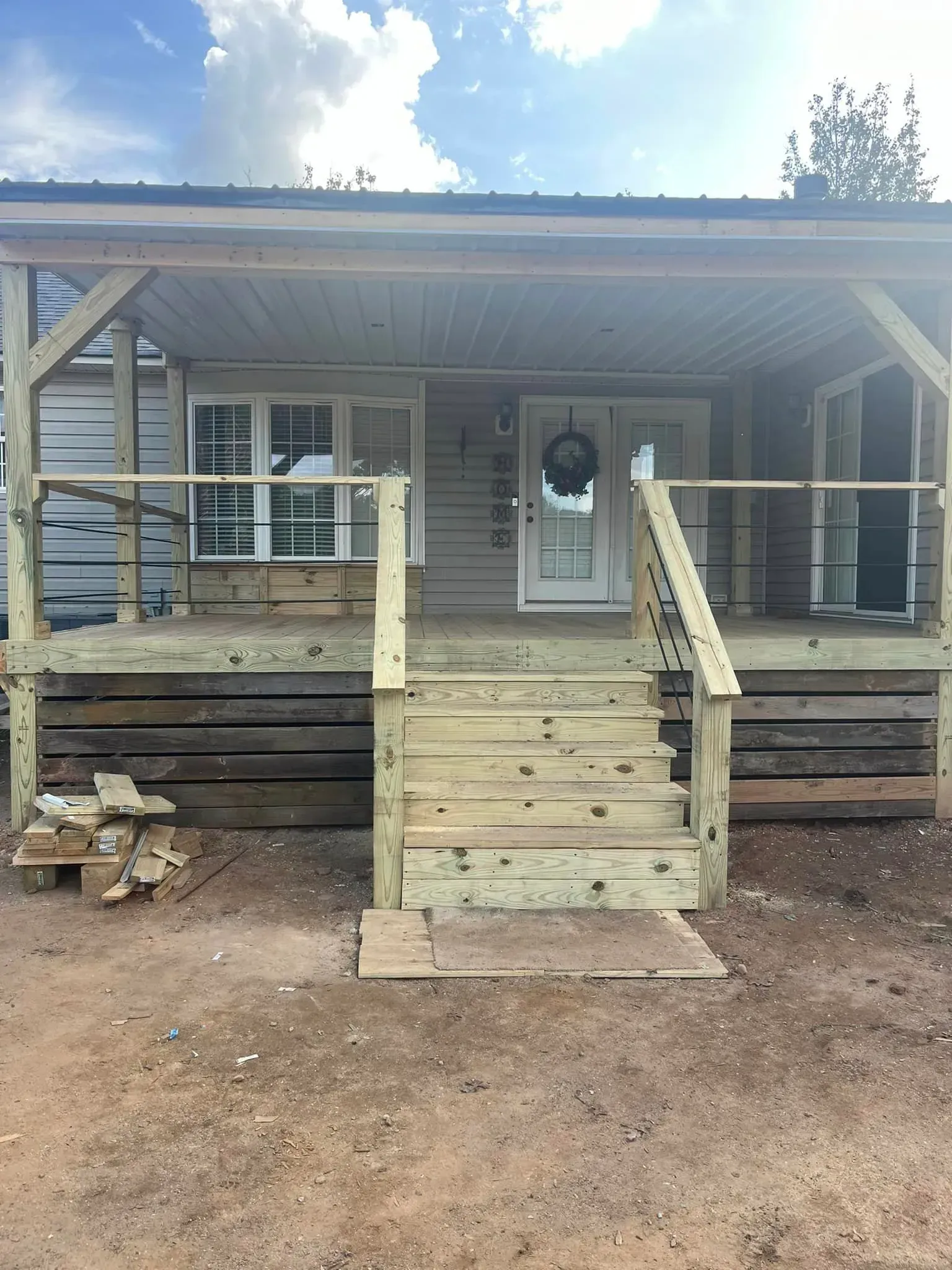 Newly built wooden porch with steps leading to a front door, set on a dirt ground, blue sky in the background.