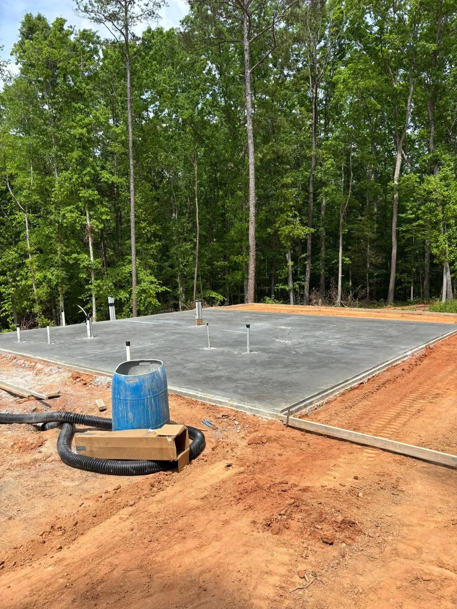 Concrete foundation with pipes in front of a treeline. Blue barrel and hoses are visible.