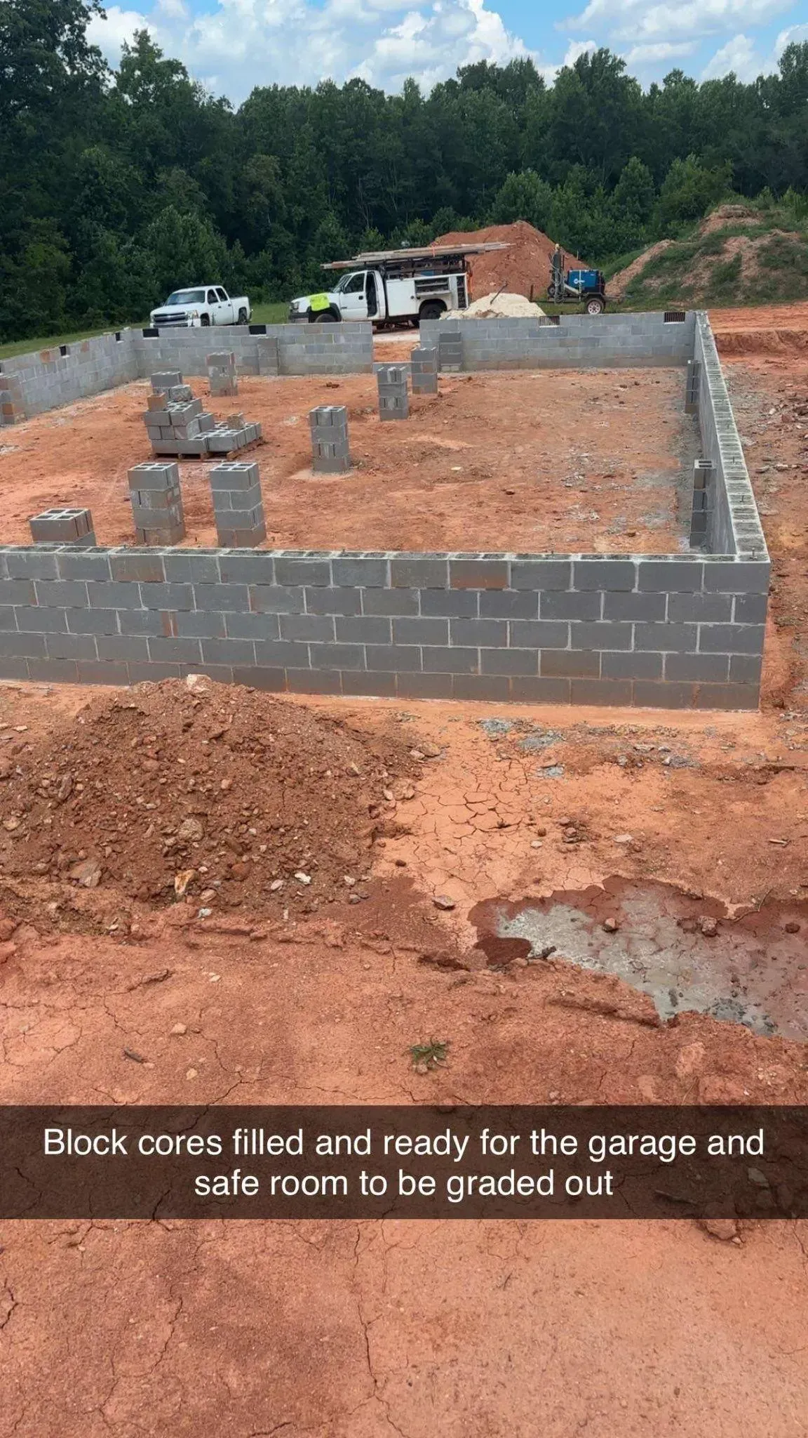 Foundation of a building under construction with block walls. Dirt, trucks, and trees in the background.