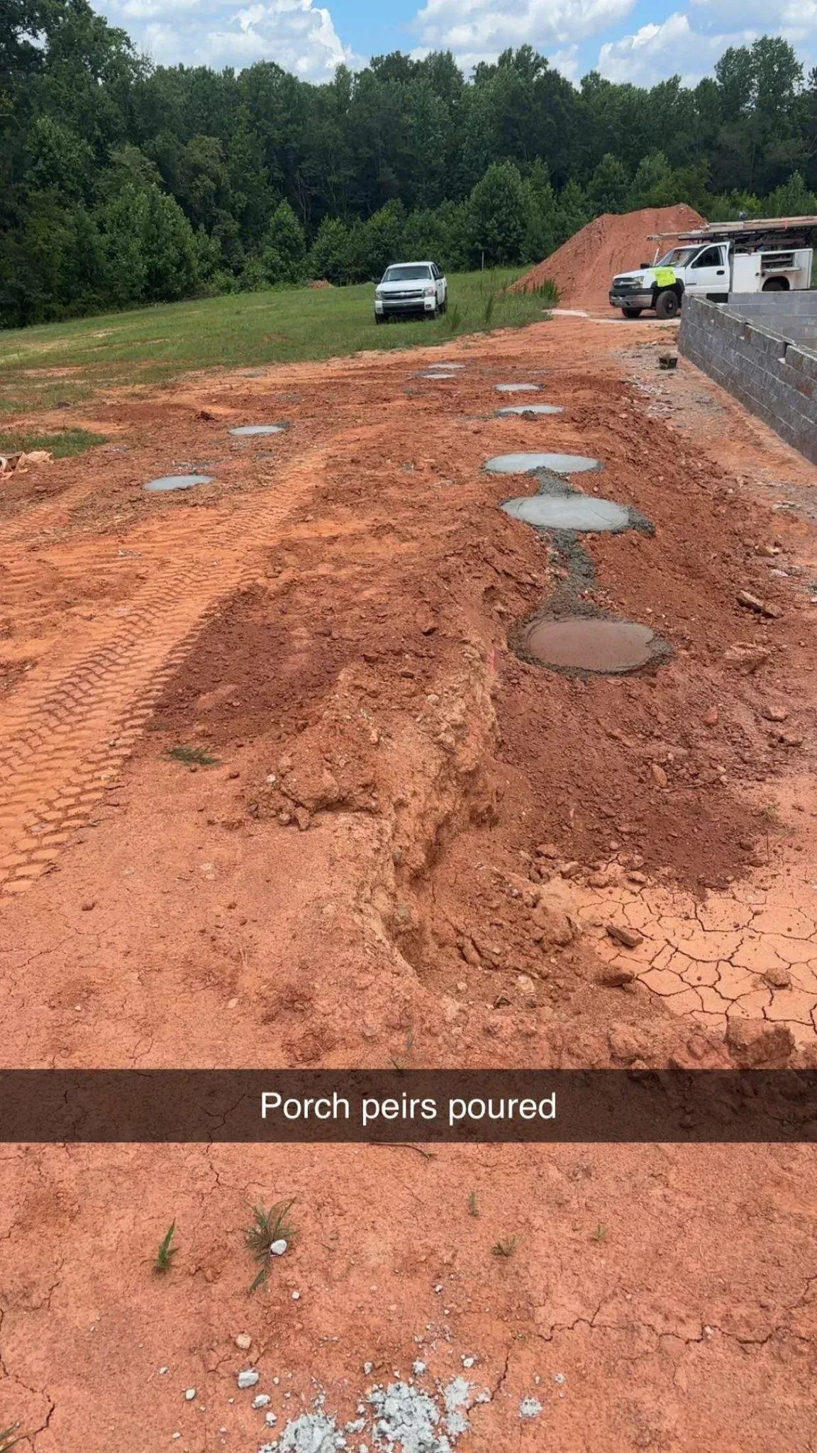 Construction site with red dirt and concrete pads. White car, trees, blue sky.
