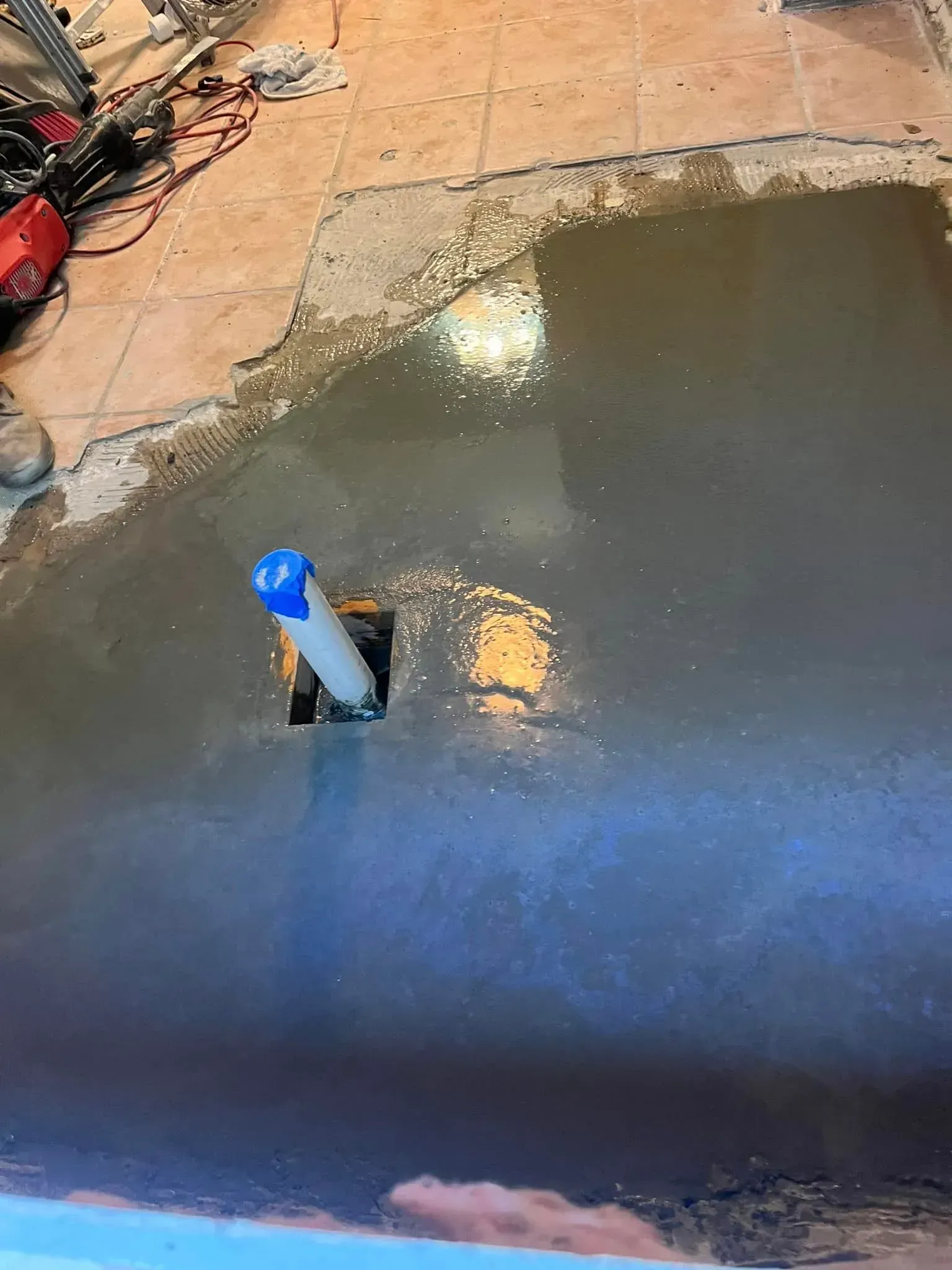 Gray cement covers a floor with exposed plumbing. A white pipe with a blue cap rises from the center.