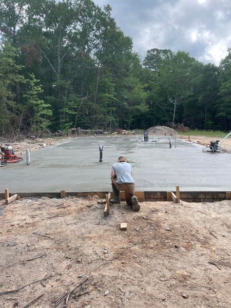Man kneels on a construction site, working on a freshly poured concrete foundation.