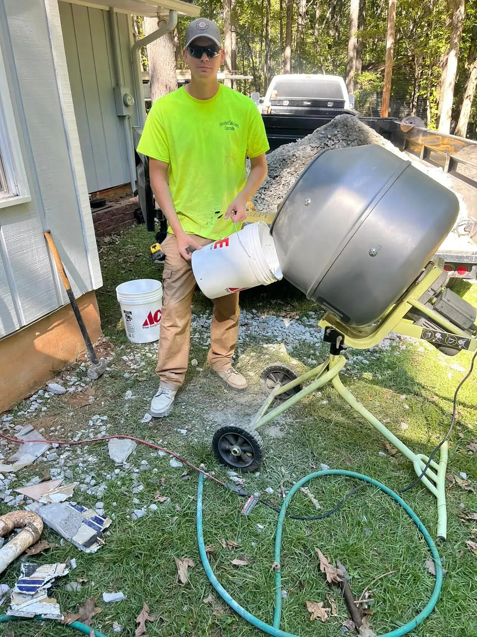 Person in neon yellow shirt holding a bucket, by a cement mixer outdoors.