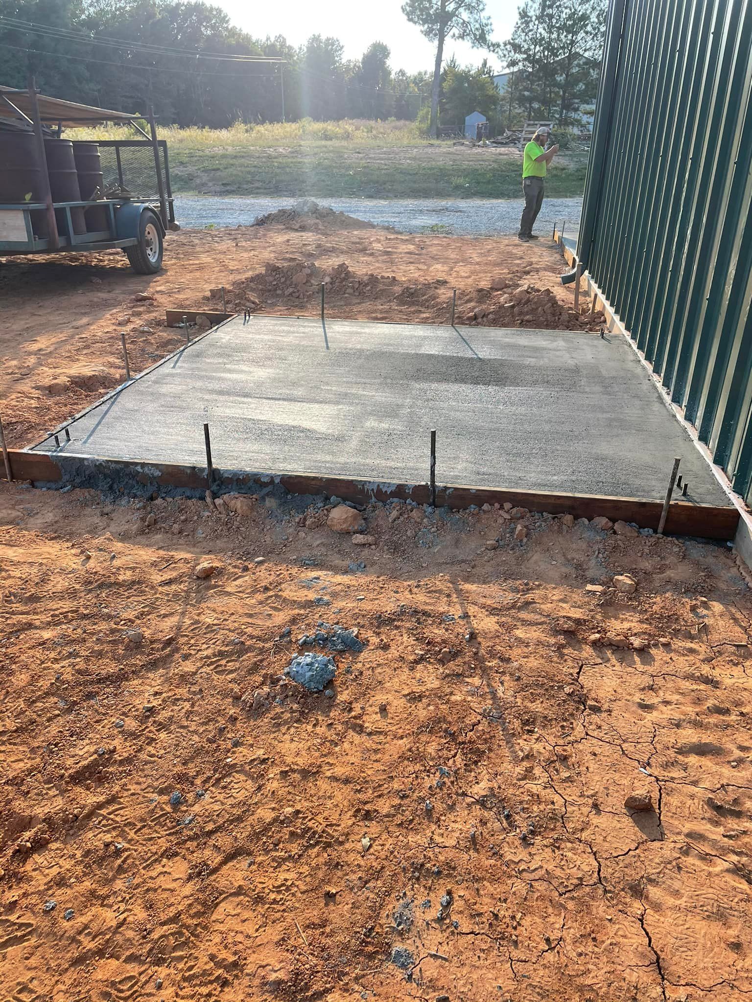 A newly poured concrete slab with rebar stakes surrounded by dirt, next to a green fence.