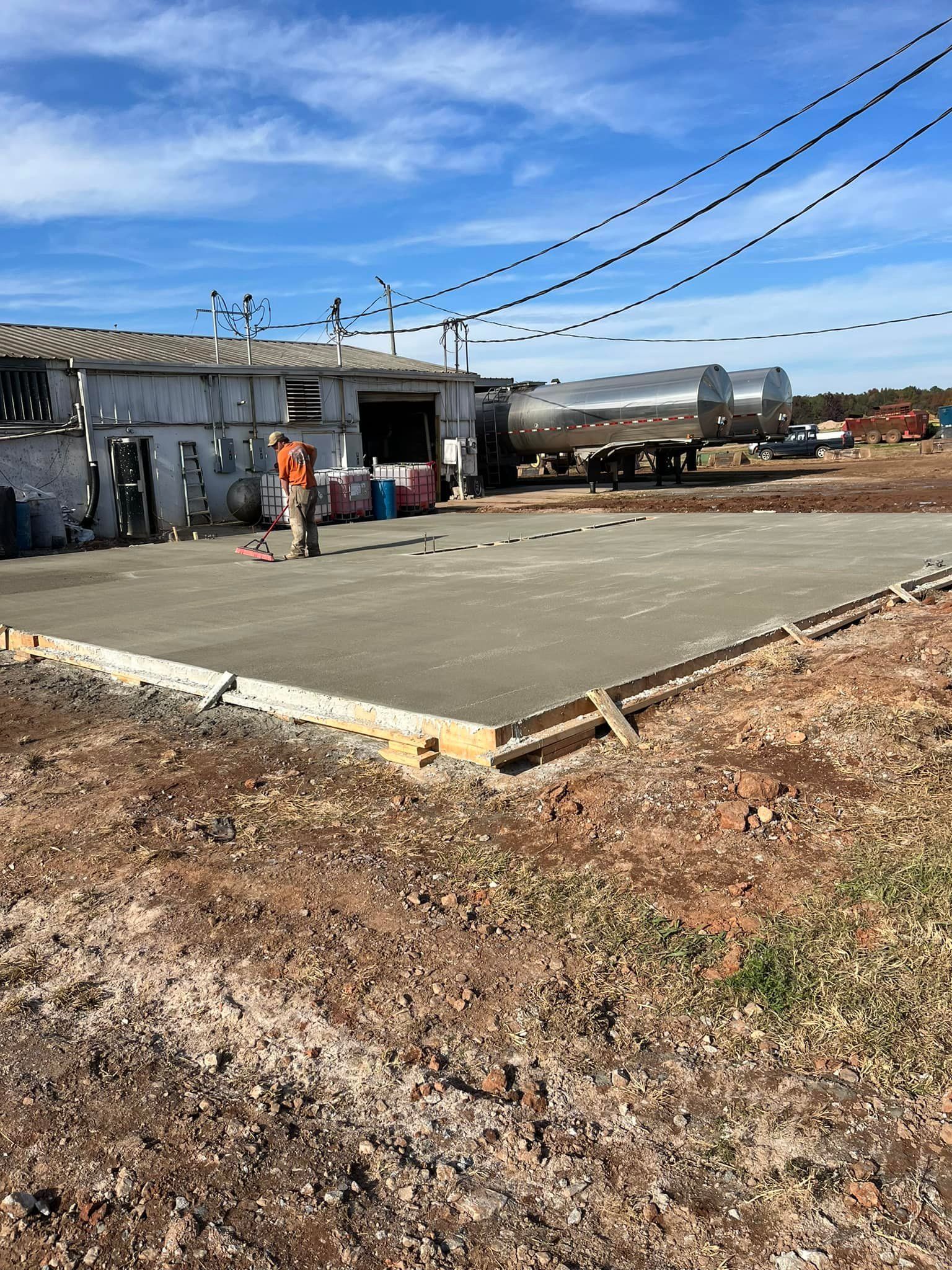 Newly poured concrete slab in front of a building; worker and tanker truck visible; blue sky overhead.