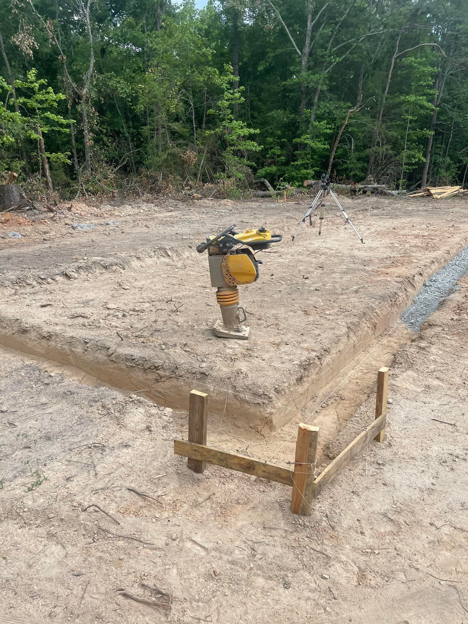 Construction site with a compactor in use, outline of foundation marked in the dirt, wooded background.