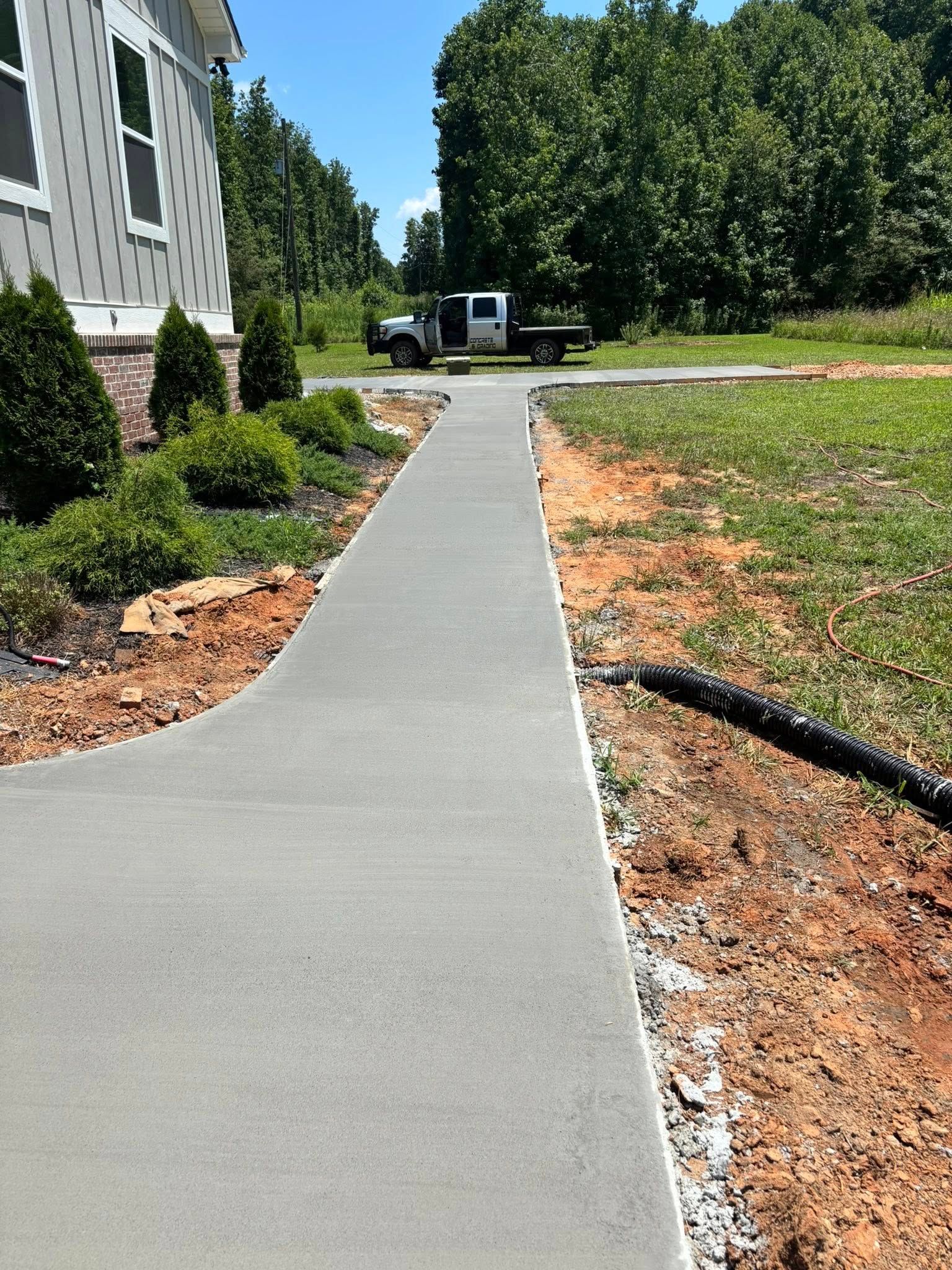 Concrete walkway leading from a house under construction to a road, with a truck parked in the distance.