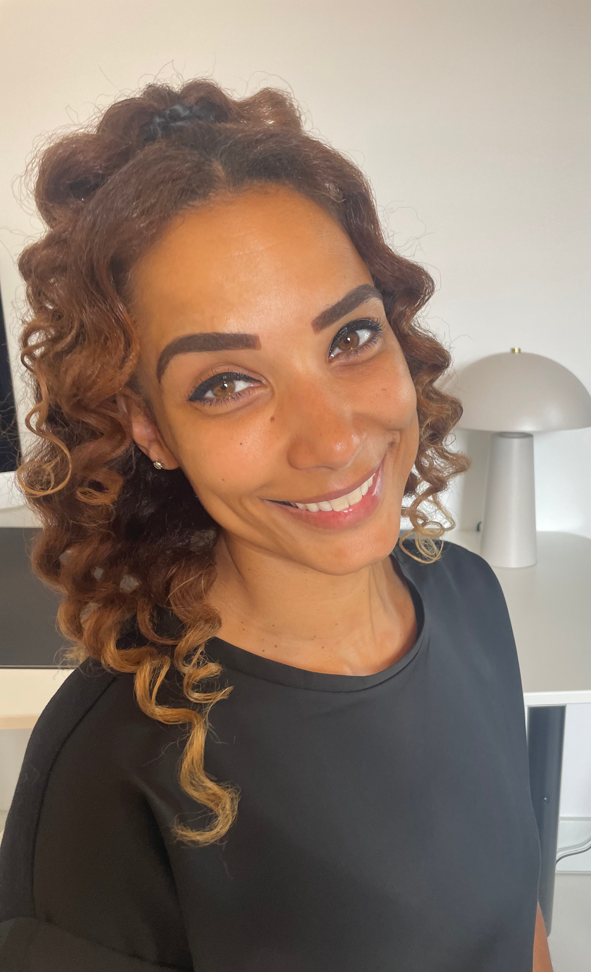 Foto vanFinch Logopediepraktijk, Een vrouw met krullend bruin haar lacht naar de camera. Ze draagt āāeen zwarte blouse en staat in een kamer met een lamp en een wit bureau.