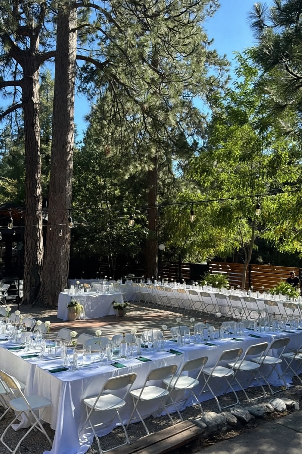 Outdoor wedding reception setup with long tables, white chairs, and trees.
