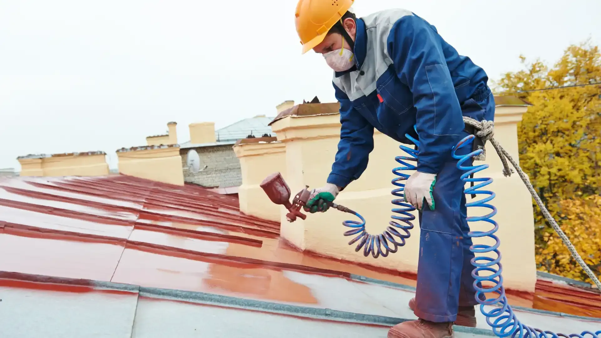 A worker in a helmet and mask sprays red paint onto a roof.
