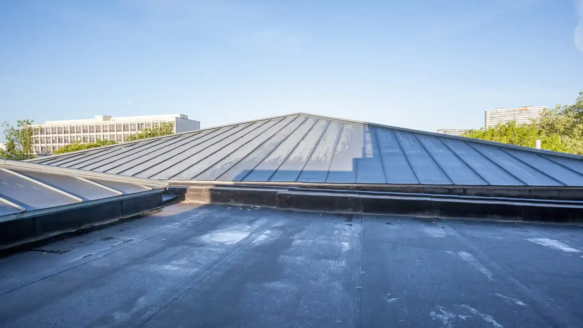 View of a dark flat roof with a peaked metal roof in the background on a sunny day.