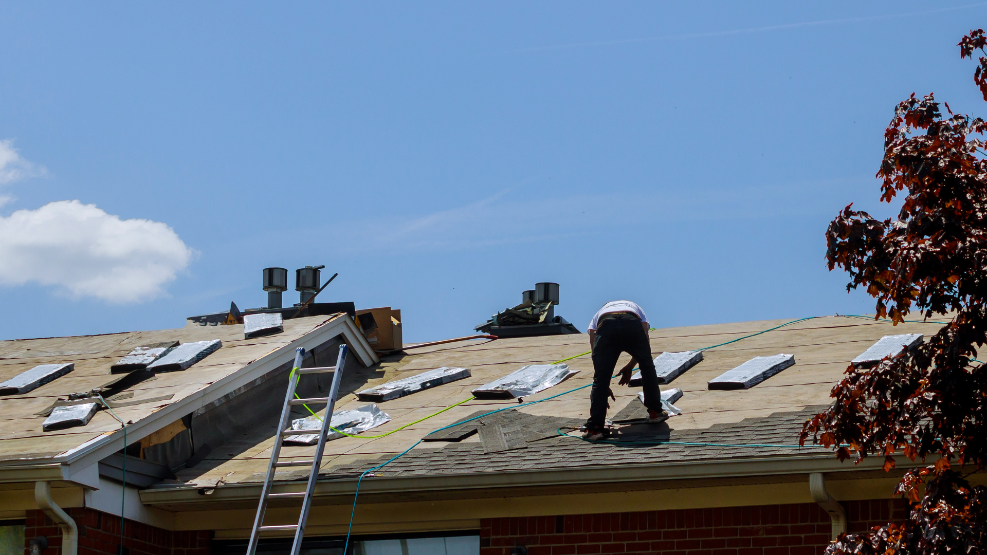 A worker in dark clothing performs repairs on a residential roof under a clear blue sky.