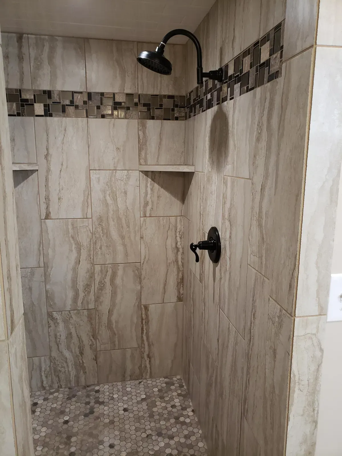 Shower with beige tiled walls, dark fixtures, mosaic tile accent, and built-in shelves.