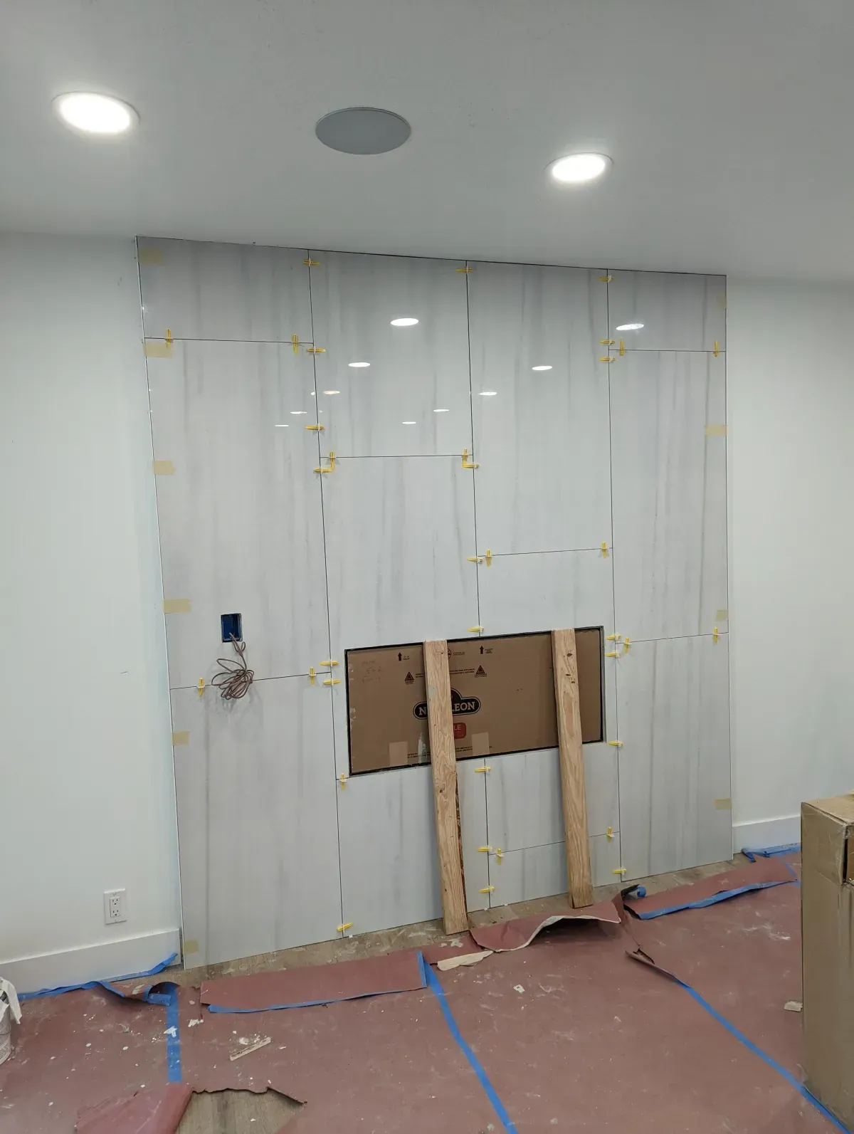 Fireplace under construction, tiled with light-colored, vertically-oriented slabs; cardboard box and wooden supports present.